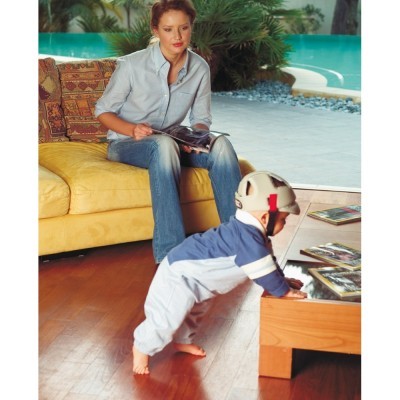 Criança com capacete e fato azul a apoiar-se numa mesa, mulher sentada num sofá amarelo lendo, ambiente interior com móveis de madeira e almofadas estampadas.