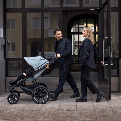 Homem e mulher a caminhar com carrinho de bebé cinzento em frente a edifício