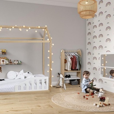 Quarto infantil com cama casinha, papel de parede arco-íris e criança sentada em tapete redondo