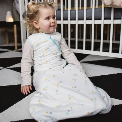 Criança sentada em tapete preto e branco usando saco de dormir branco com estrelas coloridas.