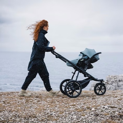 Mulher empurrando carrinho de bebé preto com capota cinzenta numa praia pedregosa