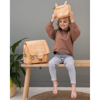 Criança com estojo e mochila bege com rosto de animal sentada numa bancada de madeira em interiores.