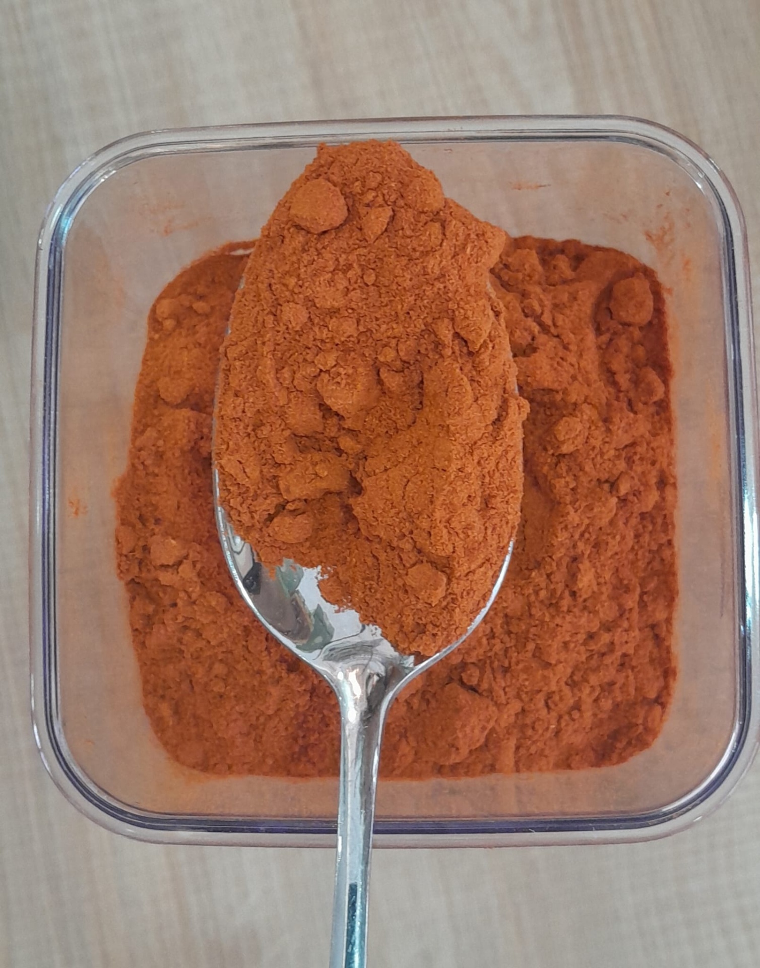 Pimenta Cayenna em Pó GRANEL Colher metálica com pó alaranjado sobre recipiente transparente com pó semelhante, colocado numa superfície de madeira clara