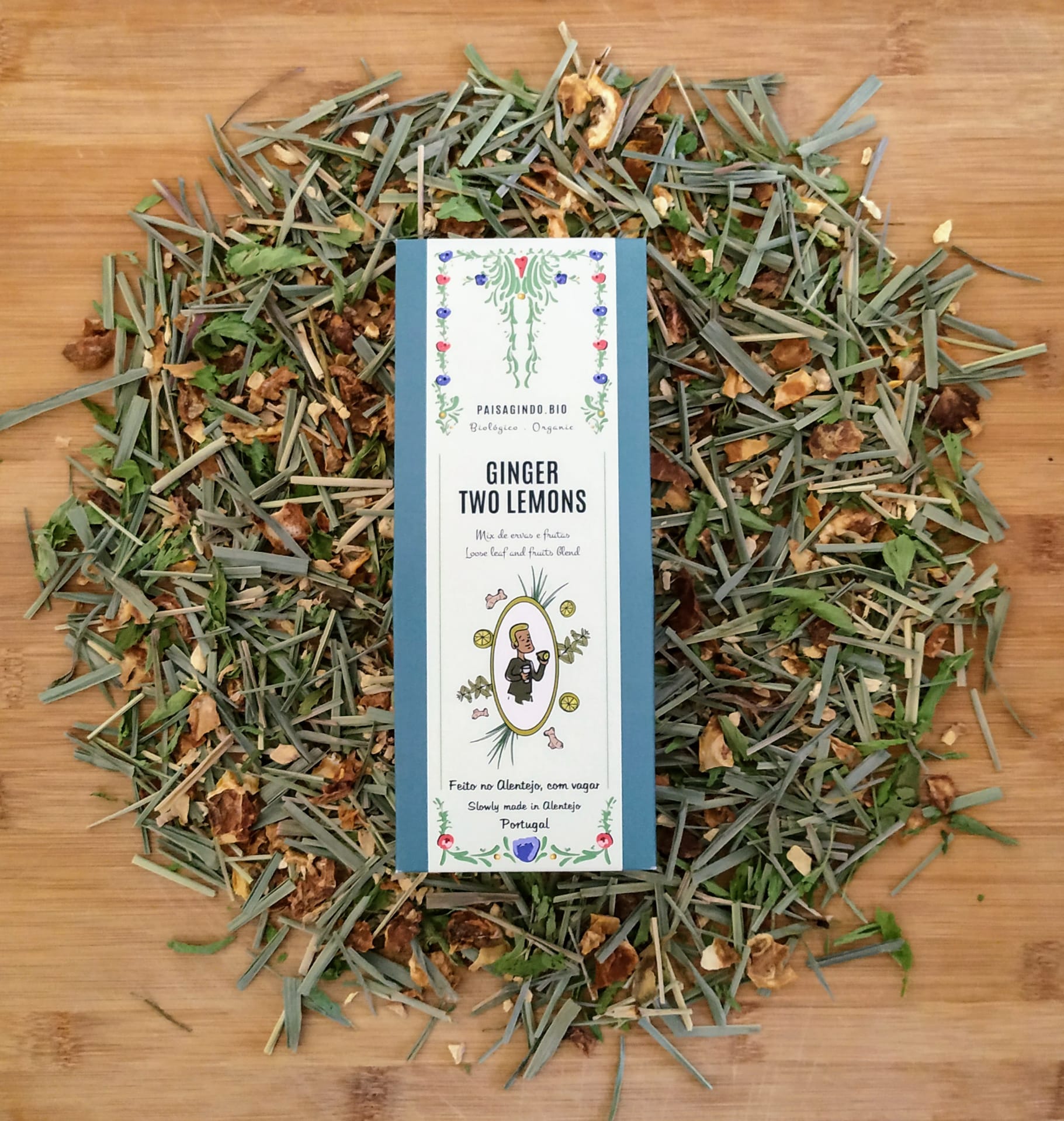Infusão Ginger Two Lemons Paisagindo 40g Pacote branco de infusão GINGER TWO LEMONS rodeado por ervas secas sobre madeira