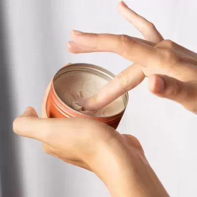 Frasco redondo laranja com creme bege claro e mão a retirar creme com o dedo