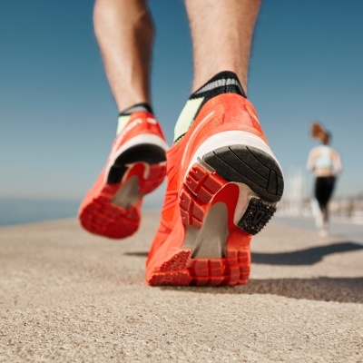 Ténis de corrida laranja com sola preta e branca e detalhes verdes numa corrida ao ar livre junto ao mar.