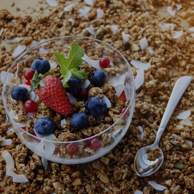Taça de vidro com granola, frutas frescas e folhas de hortelã sobre granola espalhada.