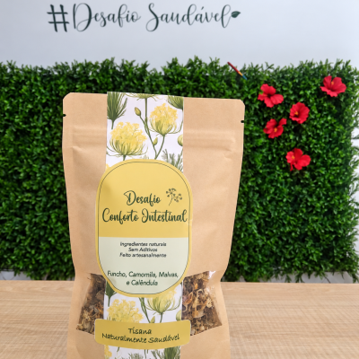 Embalagem de tisana natural com rótulo amarelo e branco, sobre mesa de madeira com fundo de plantas verdes e flores vermelhas.