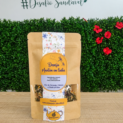 Embalagem de tisana com etiqueta amarela e branca sobre mesa de madeira e fundo com sebes e flores vermelhas
