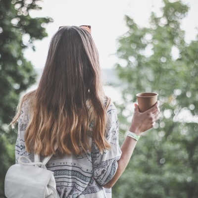 Mulher de costas com mochila branca e copo de papel, vista exterior com árvores
