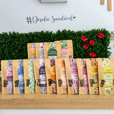 Pacotes de tisanas naturais em sacos kraft com etiquetas coloridas, expostos em mesa de madeira com plantas e texto #Desafio Saudável ao fundo