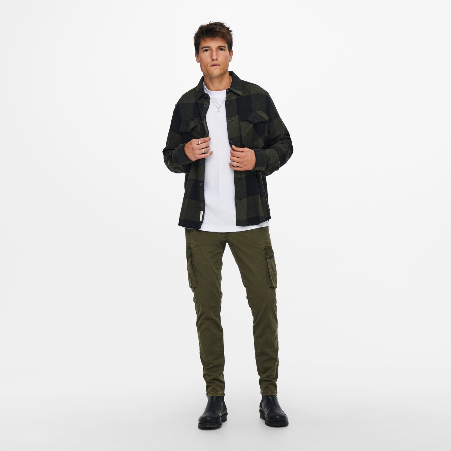 Homem a usar camisa xadrez preta e verde, t-shirt branca, calças cargo verdes e botas pretas