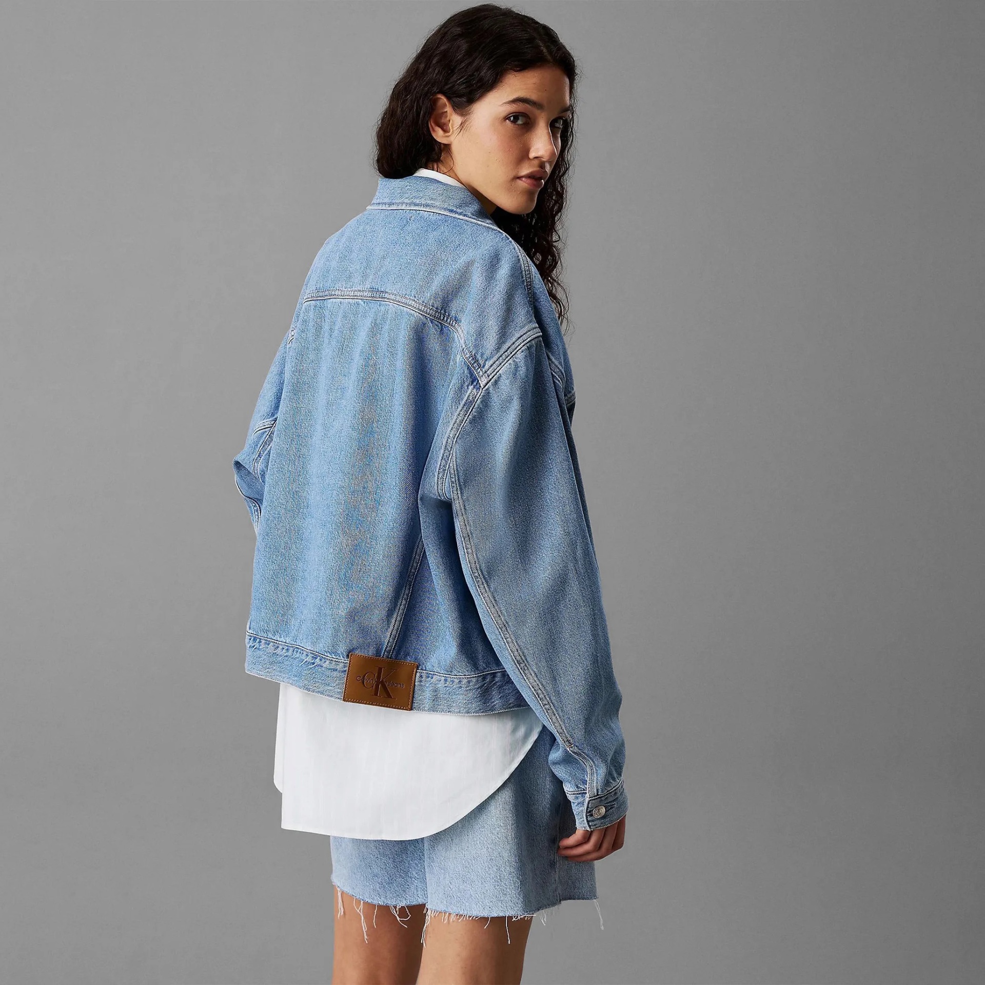 Casaco de ganga azul claro com etiqueta CALVIN KLEIN JEANS nas costas, usado com camisa branca e calções de ganga azul claro.