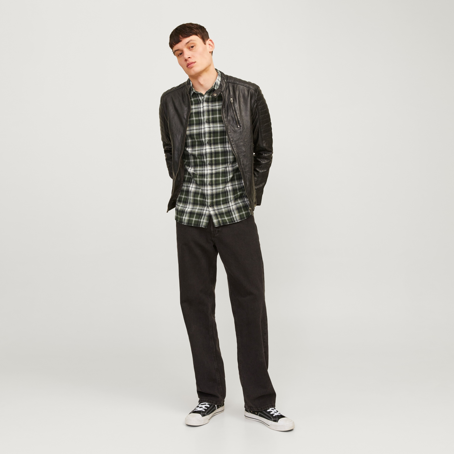 Homem com blusão em pele preta, camisa xadrez verde e calças pretas largas