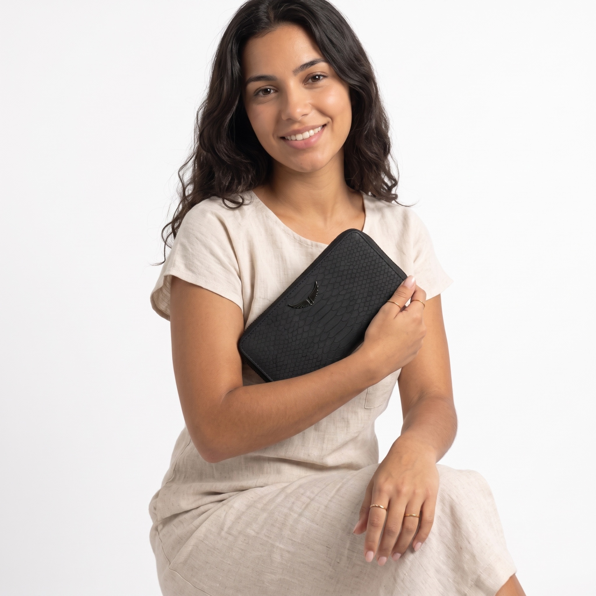 Mulher sorridente com vestido bege segurando carteira preta com padrão de escamas