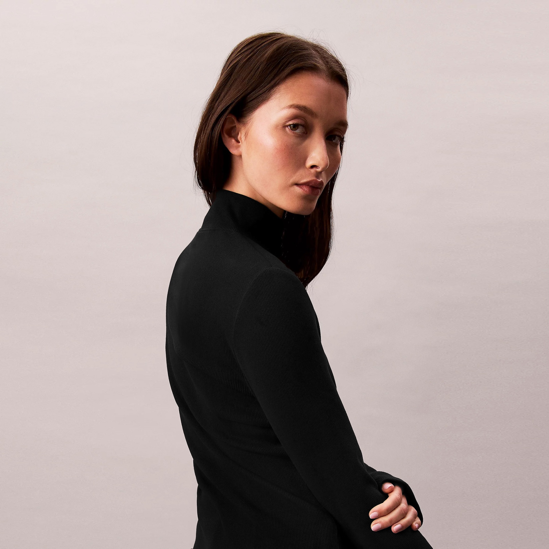 Mulher com vestido preto de malha de gola alta num fundo cinza-claro