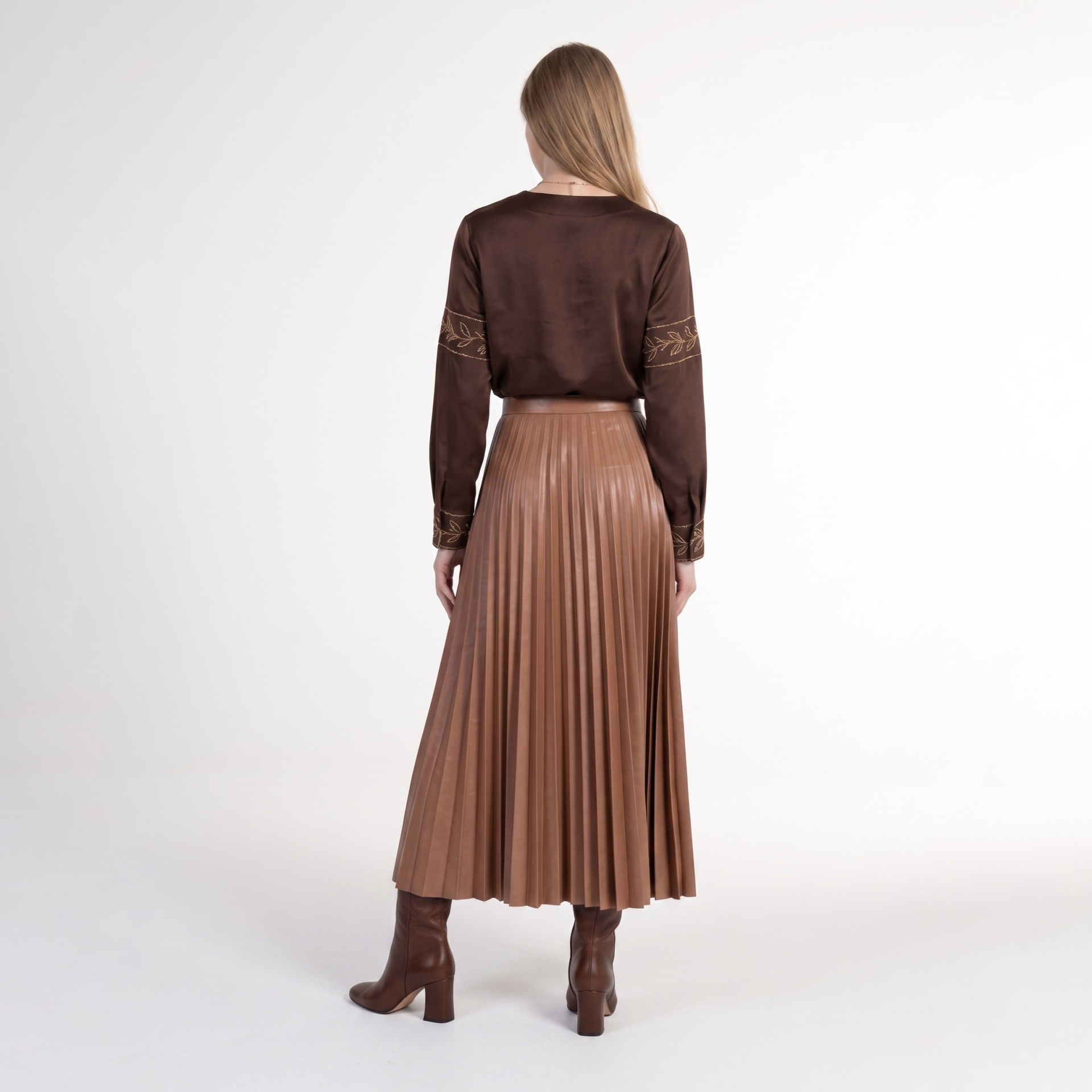 Mulher de costas com blusa castanha acetinada com bordados e saia plissada castanha com botas castanhas.