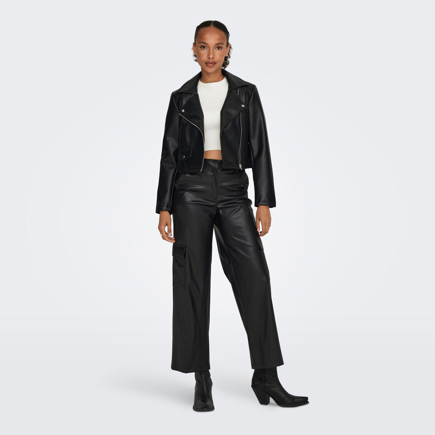 mulher com casaco e calças pretas de pele sintética e top branco, em fundo cinzento