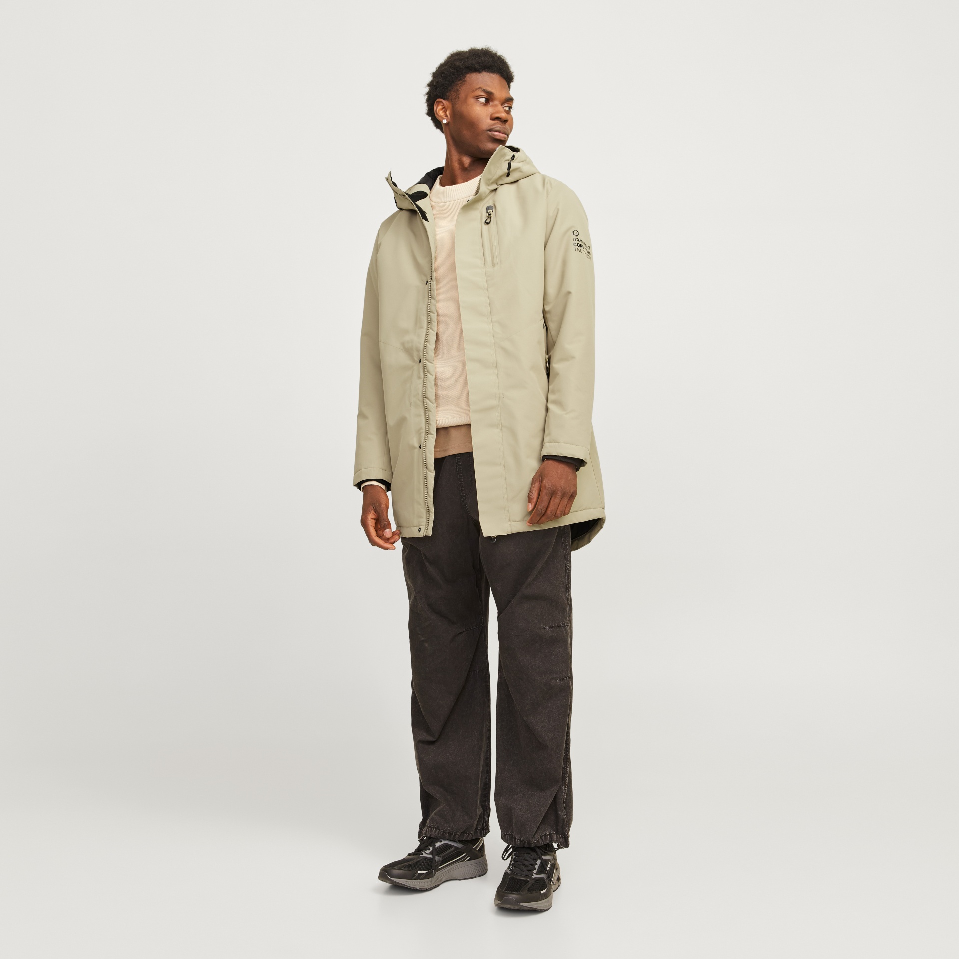 Homem com casaco bege, calças pretas e sapatilhas pretas, em fundo branco