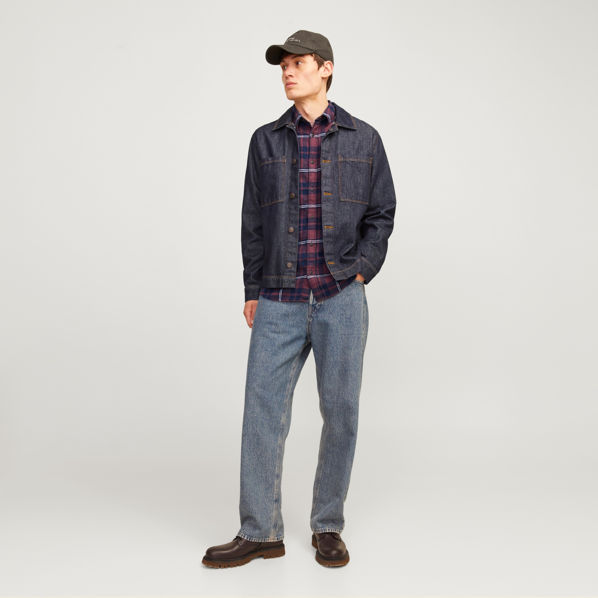 Homem com casaco azul escuro, camisa xadrez, jeans azuis, sapatos castanhos e boné cinzento escuro.