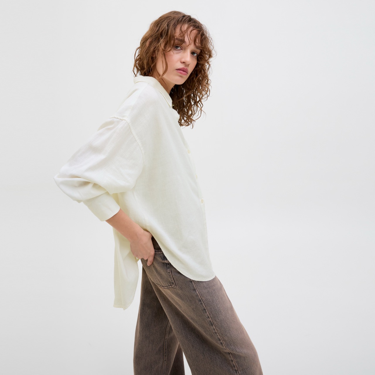 Mulher vestindo camisa branca de tecido leve e calças castanhas largas em fundo branco