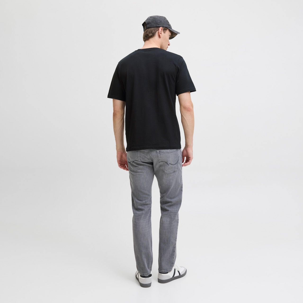 Homem de costas com boné cinzento, t-shirt preta, calças de ganga cinza e ténis brancos