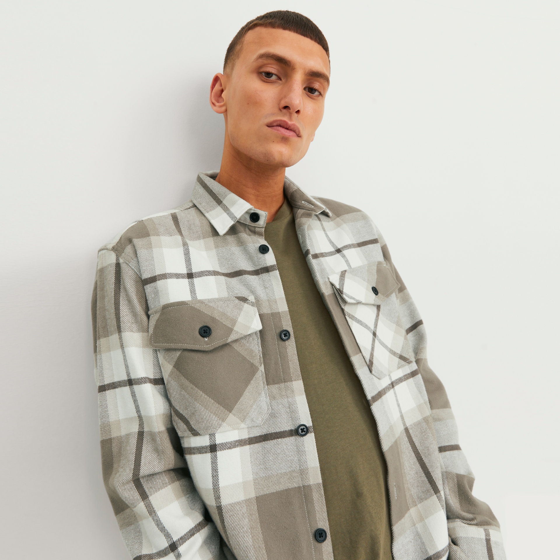 Camisa xadrez bege, branco e cinza com botões pretos e bolsos, sobre t-shirt verde oliva, com modelo masculino