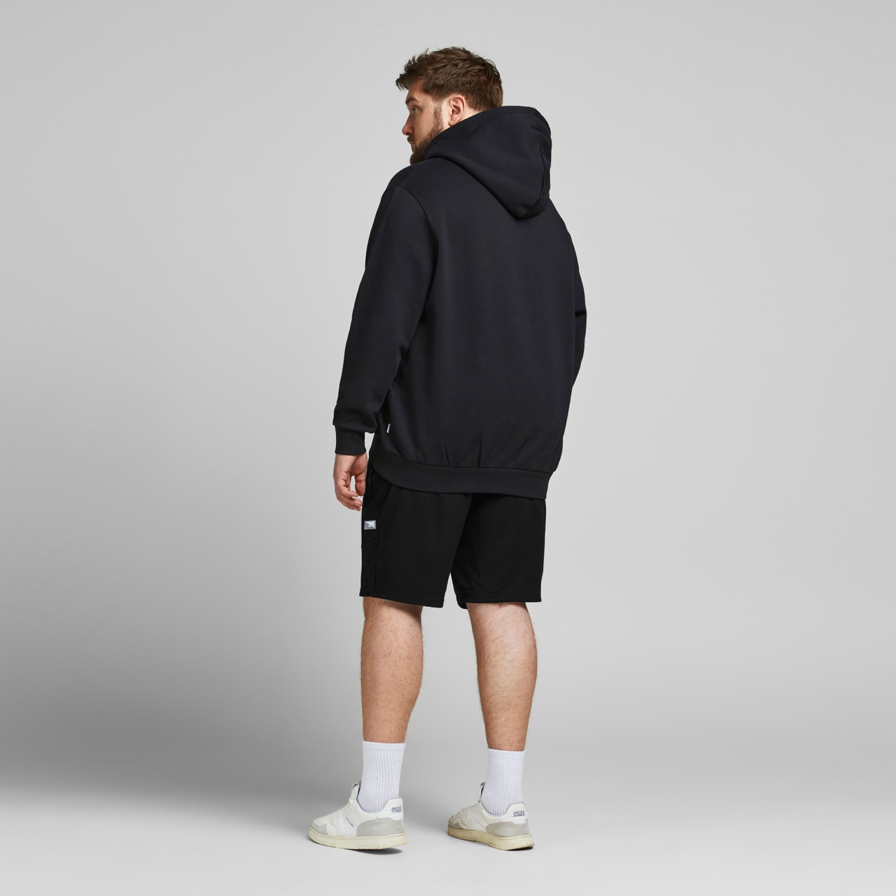 Homem de costas com hoodie e calções pretos em fundo cinzento.