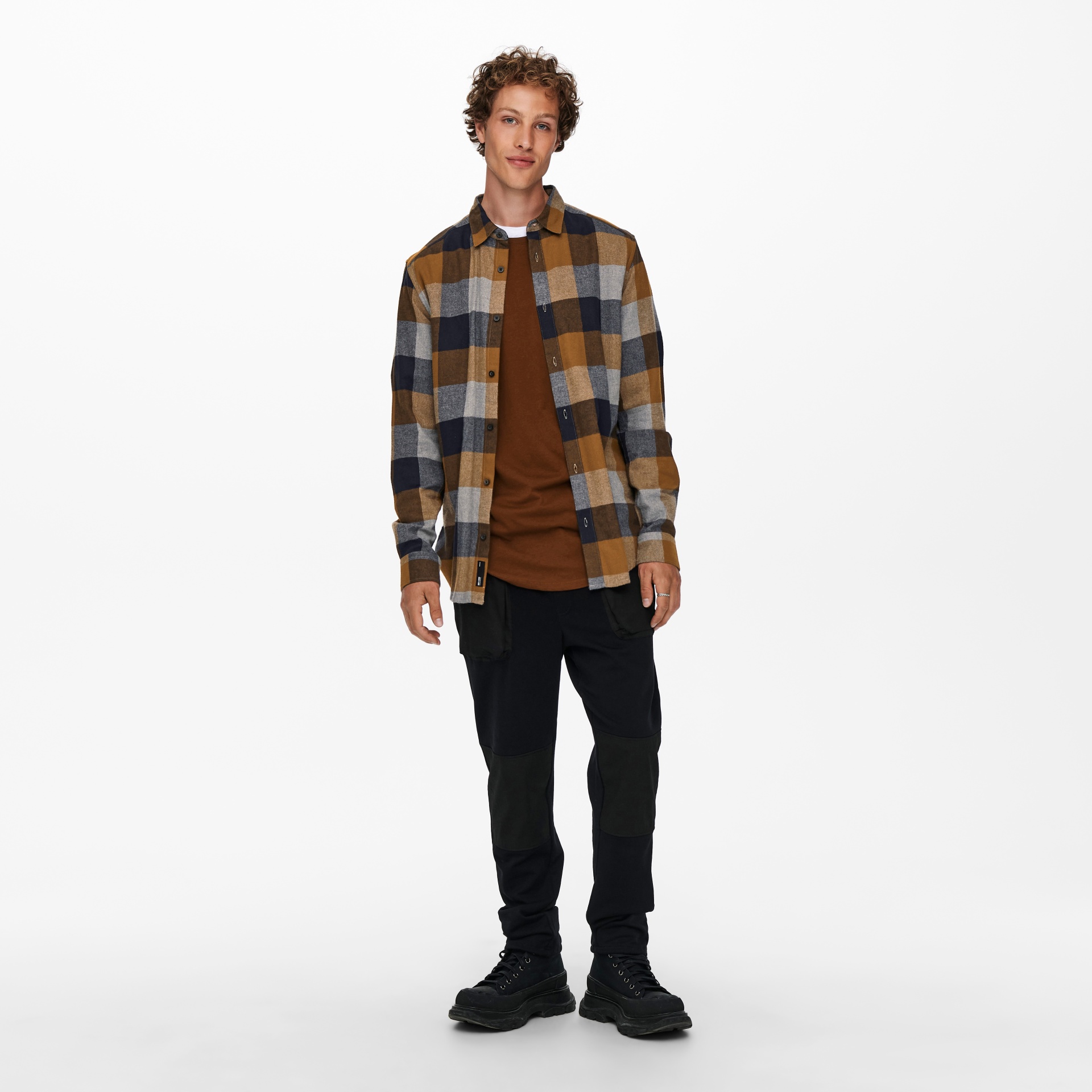 Homem com camisa xadrez castanha e cinza, t-shirt castanha, calças pretas e sapatos pretos