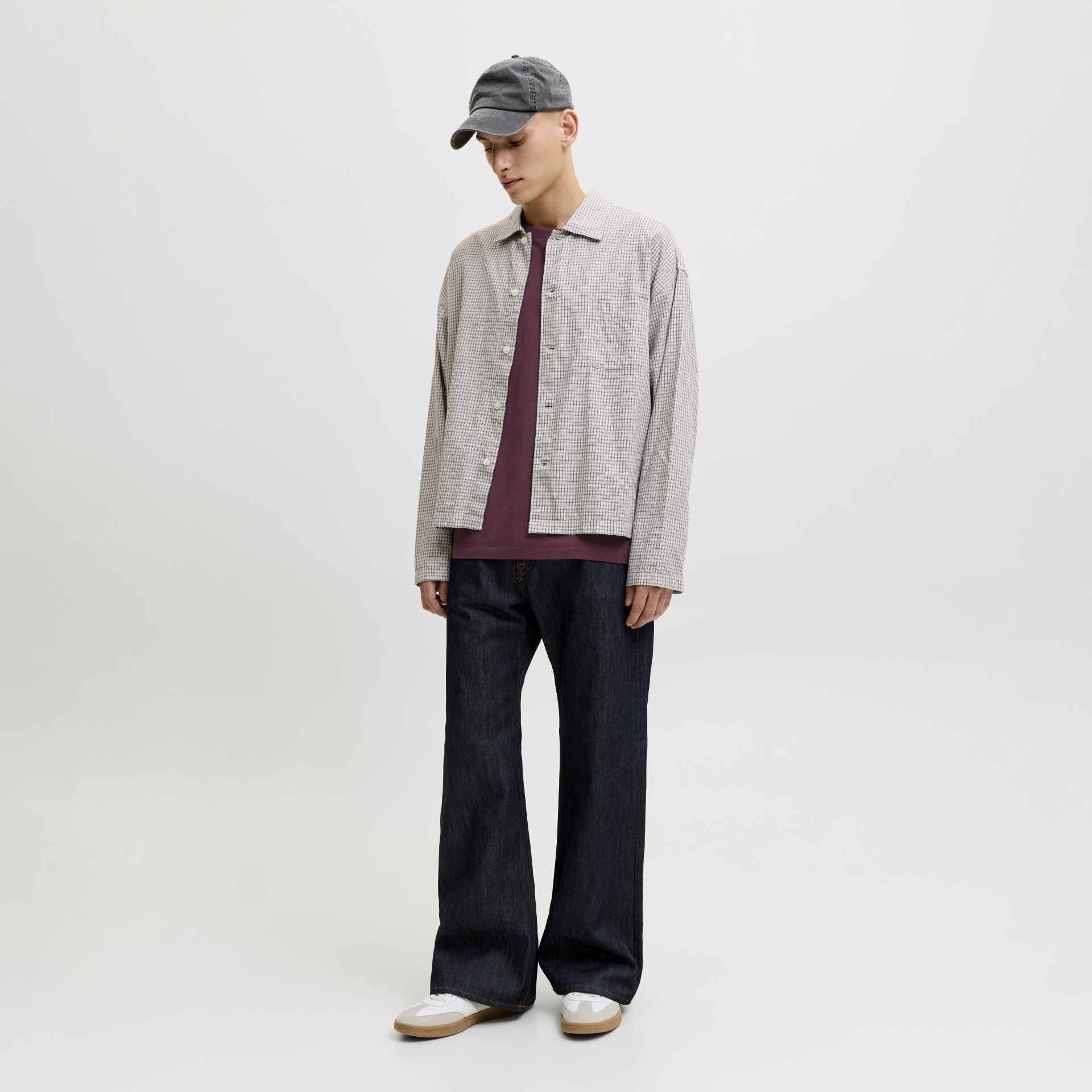 Homem com camisola xadrez bege, t-shirt roxa, calças de ganga escuras, boné cinzento e sapatilhas brancas