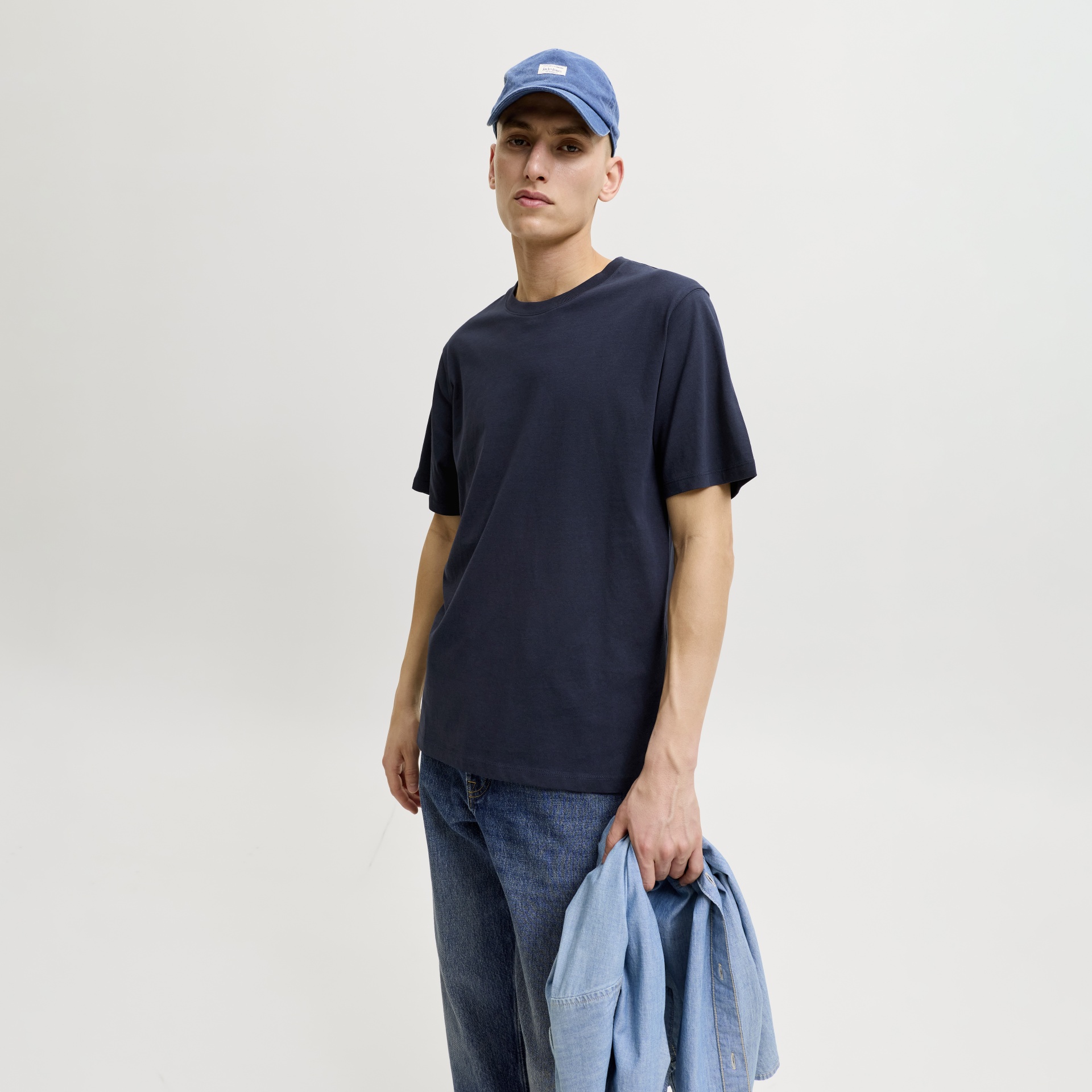 Homem com t-shirt azul escuro, boné azul e calças de ganga, segurando camisa de ganga azul claro