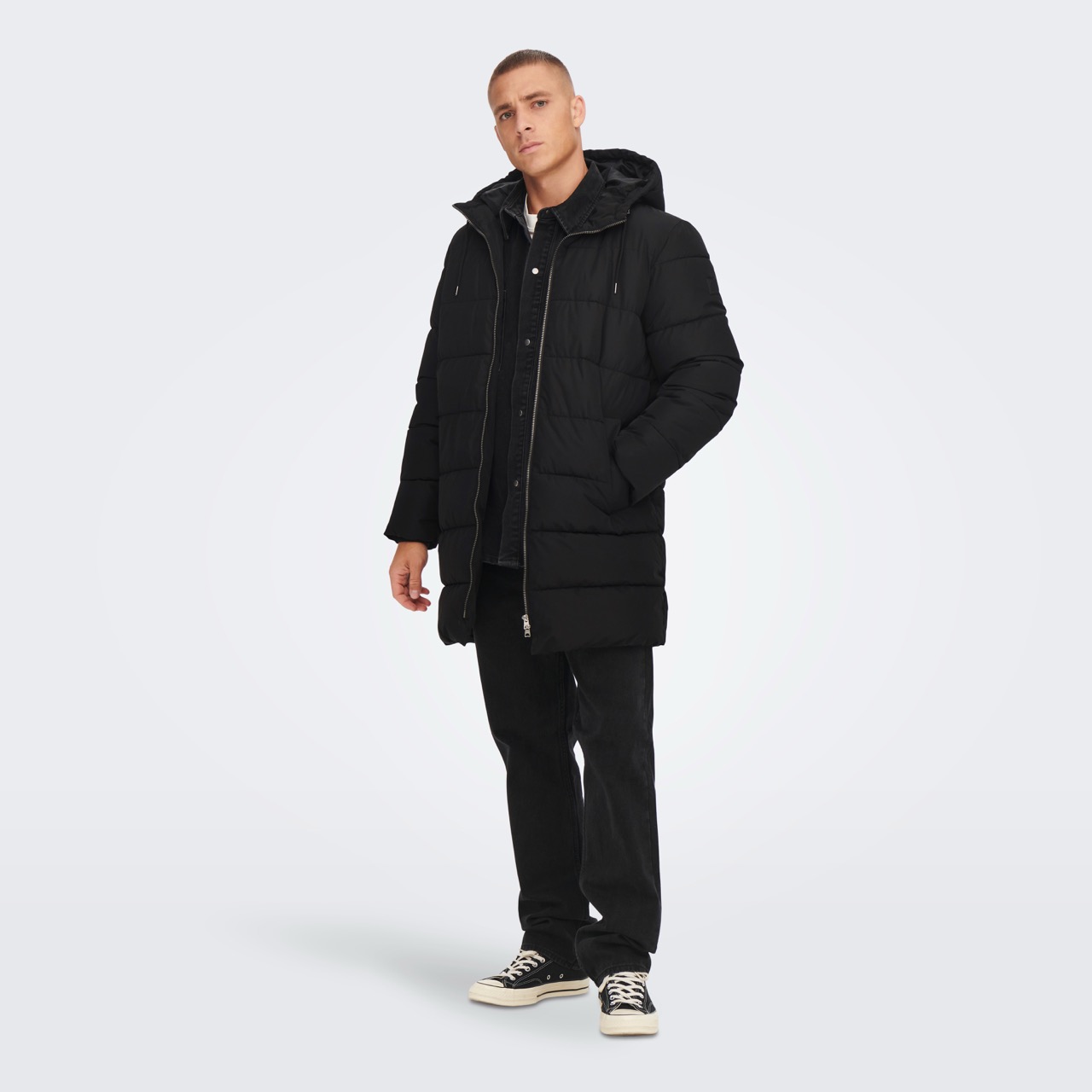 Homem de casaco acolchoado preto com capuz, camisa e jeans pretos e ténis preto e branco em fundo cinzento