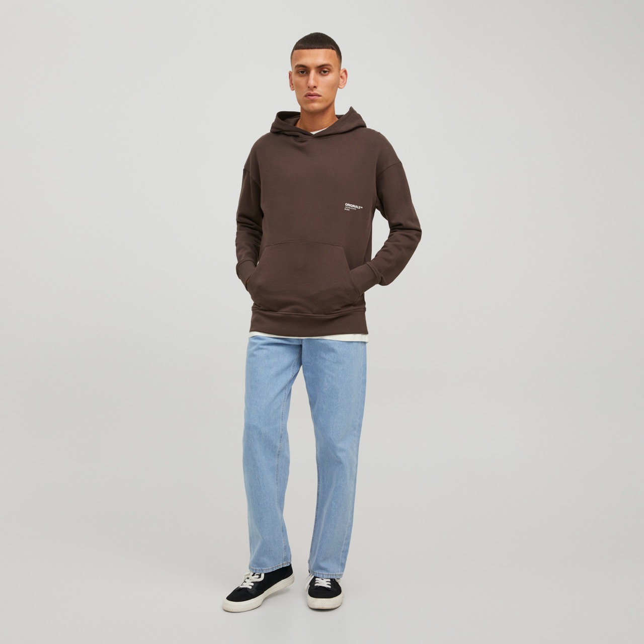 Homem a vestir hoodie castanho escuro, calças de ganga azul claro e sapatilhas pretas com branco em fundo branco