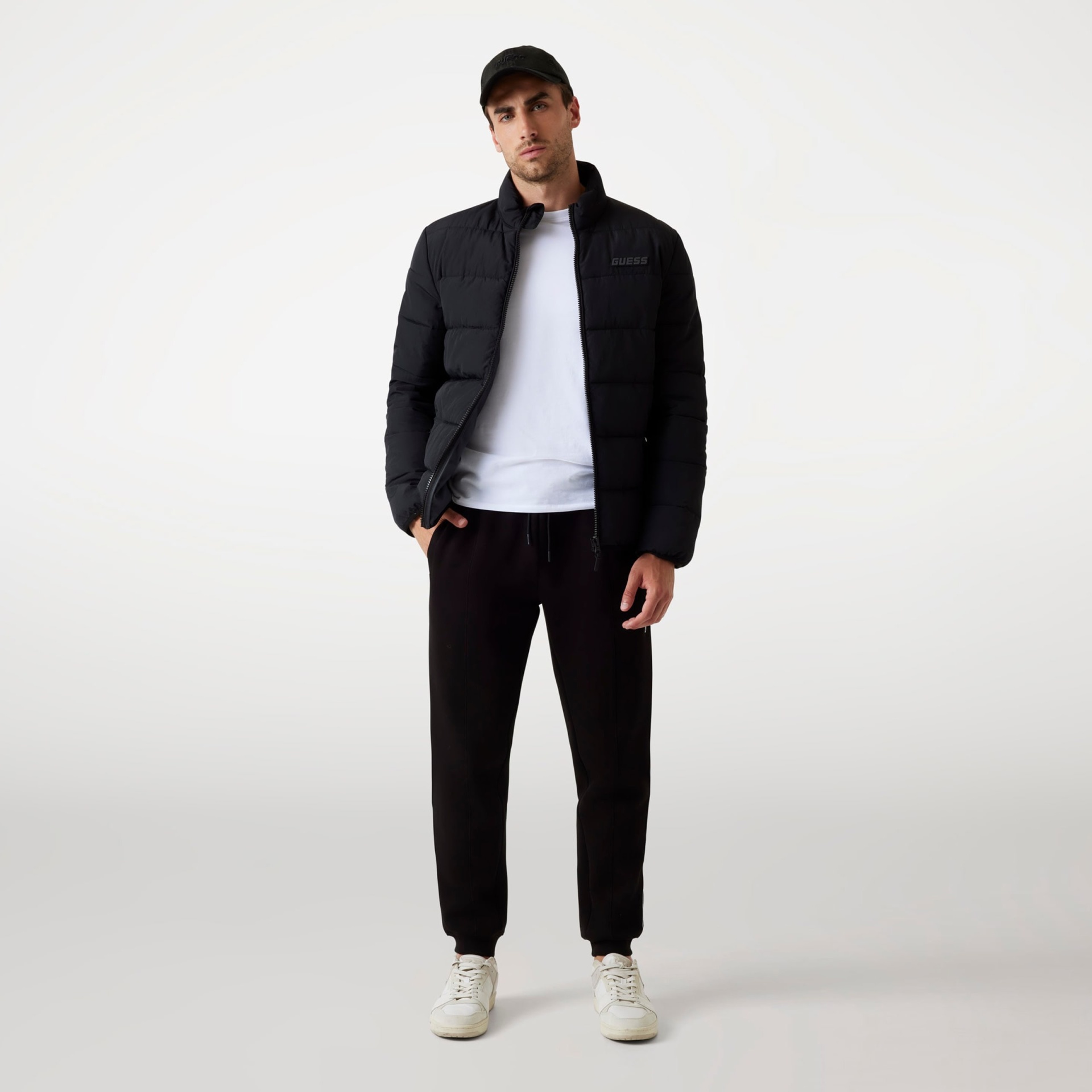 Homem de pé vestindo casaco acolchoado preto, camisola branca, calças pretas e ténis brancos