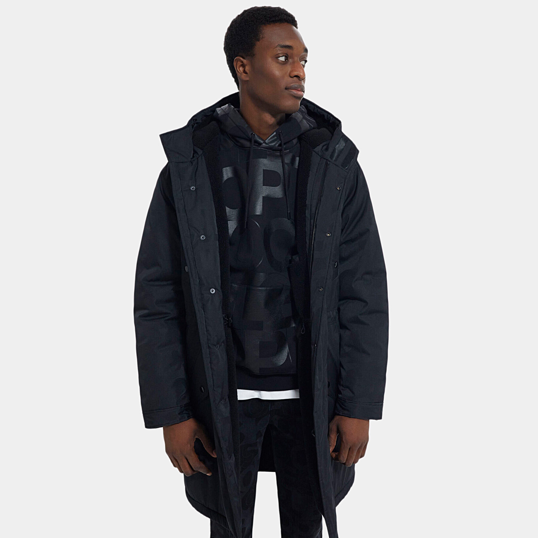 Homem com casaco comprido preto e hoodie preto com letras grandes.