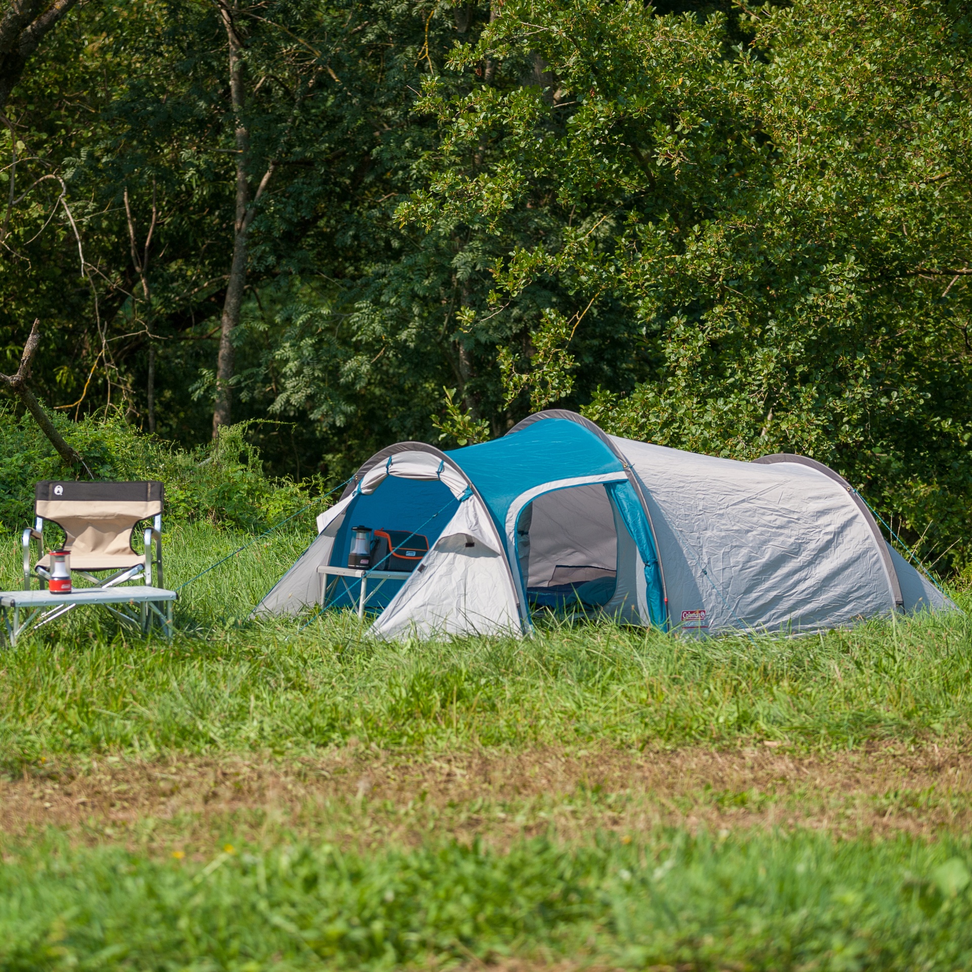 Tenda de campismo e mobiliário exterior em campo verde com árvores ao fundo