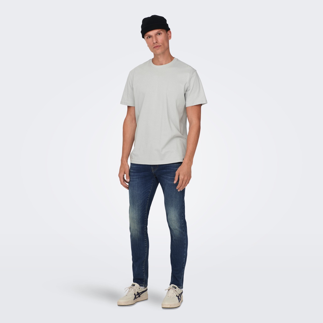 Homem com t-shirt cinza, jeans azul escuro, sapatilhas claras e gorro preto em fundo cinza claro