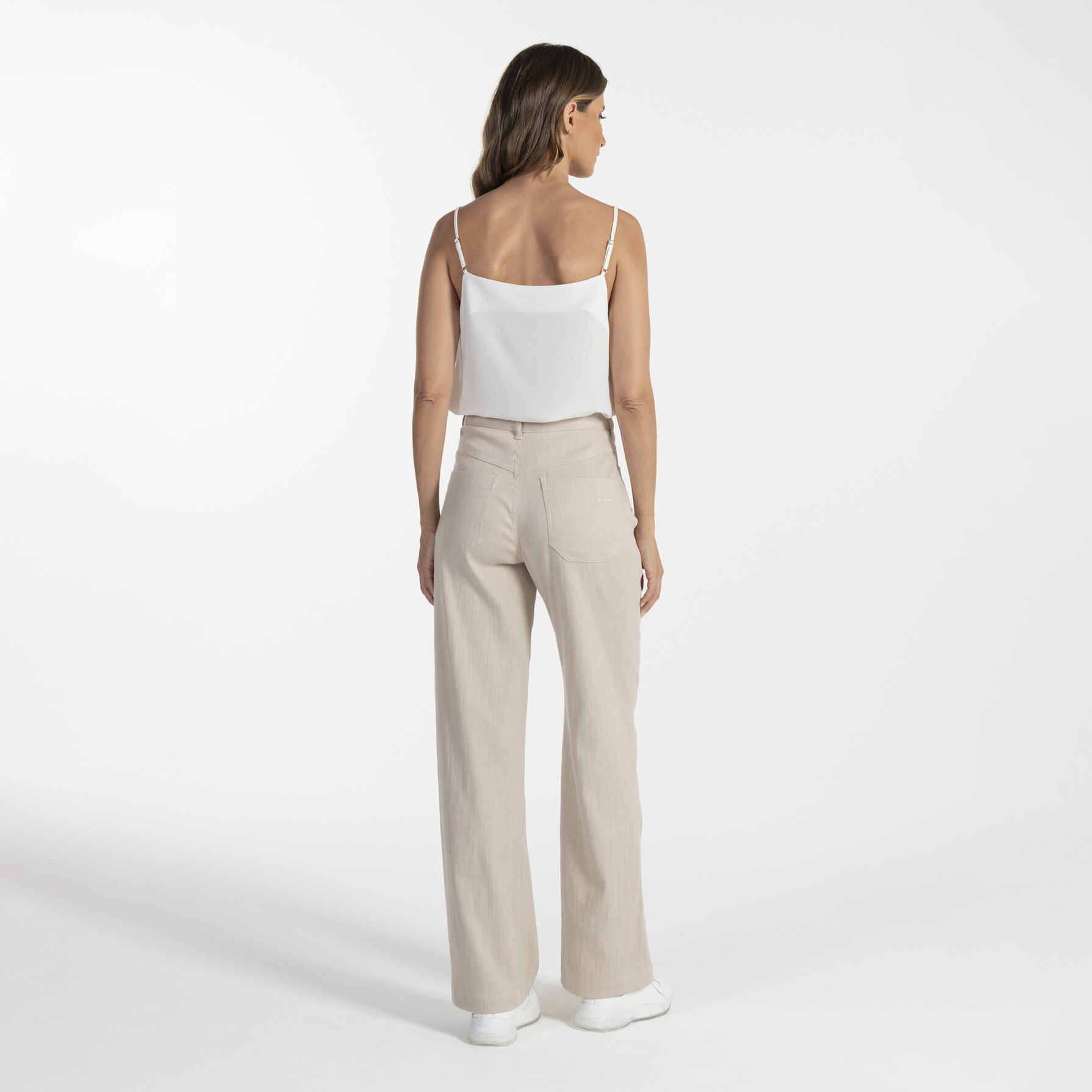 Mulher de costas com top branco e calças bege em fundo branco.