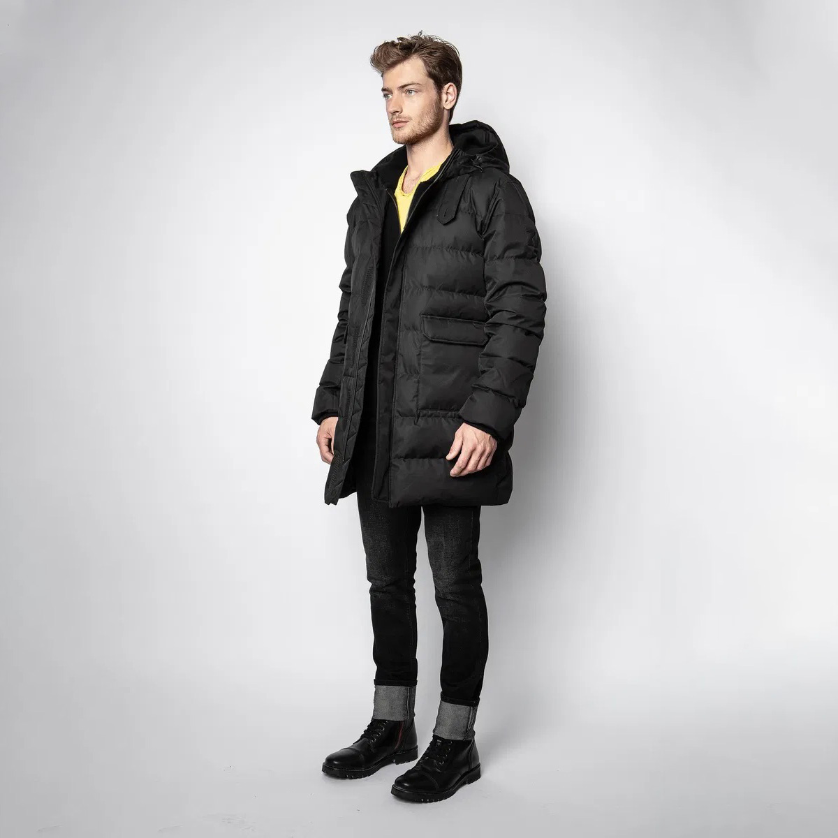 Homem com casaco preto acolchoado, capuz e botas pretas, fundo branco.