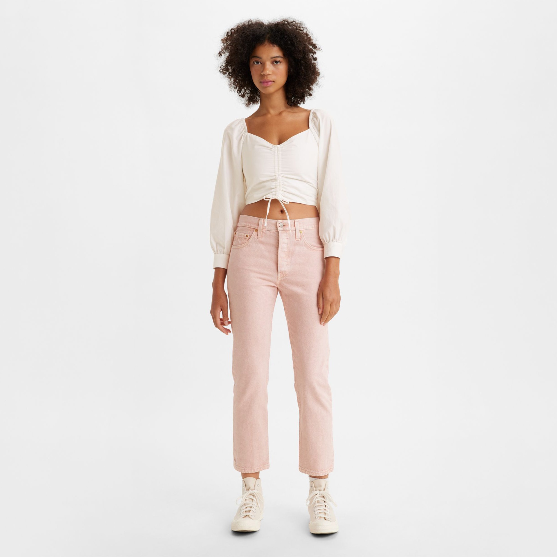 Mulher com blusa branca de manga comprida e calças rosa claro junto a fundo branco