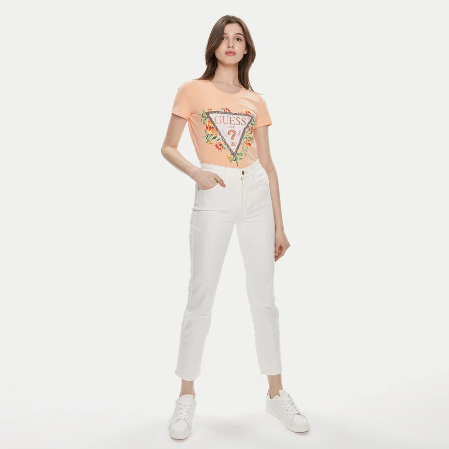 Modelo feminina com t-shirt laranja estampada Guess, calças brancas e ténis brancos em fundo branco