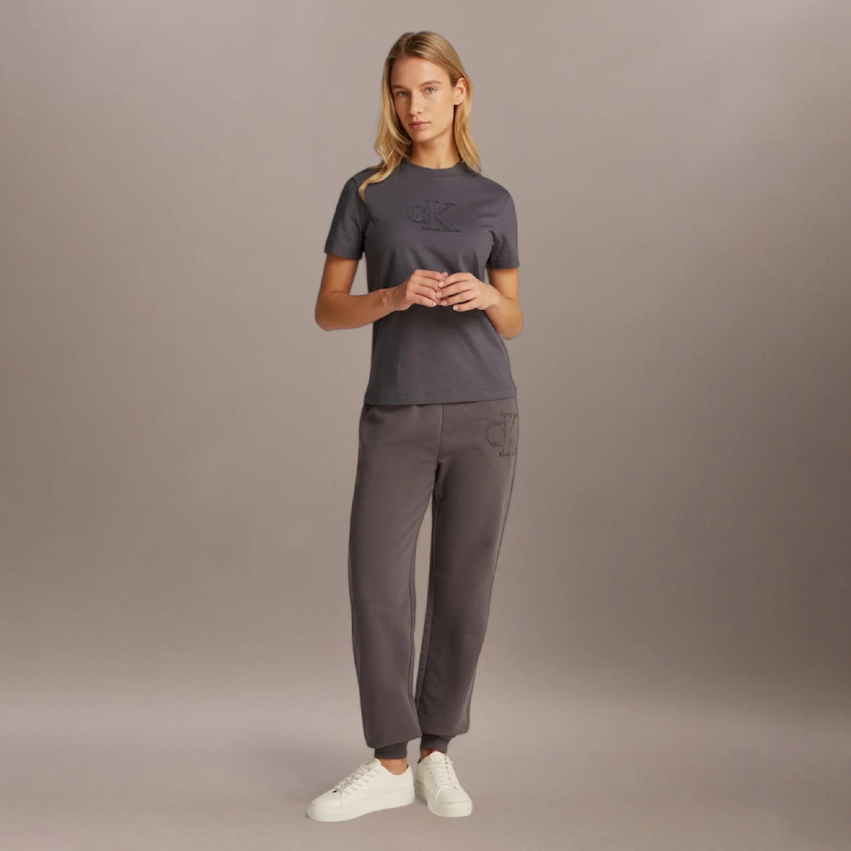 Mulher loira de pé vestindo conjunto cinzento com t-shirt e calças, sapatos brancos, fundo cinzento.
