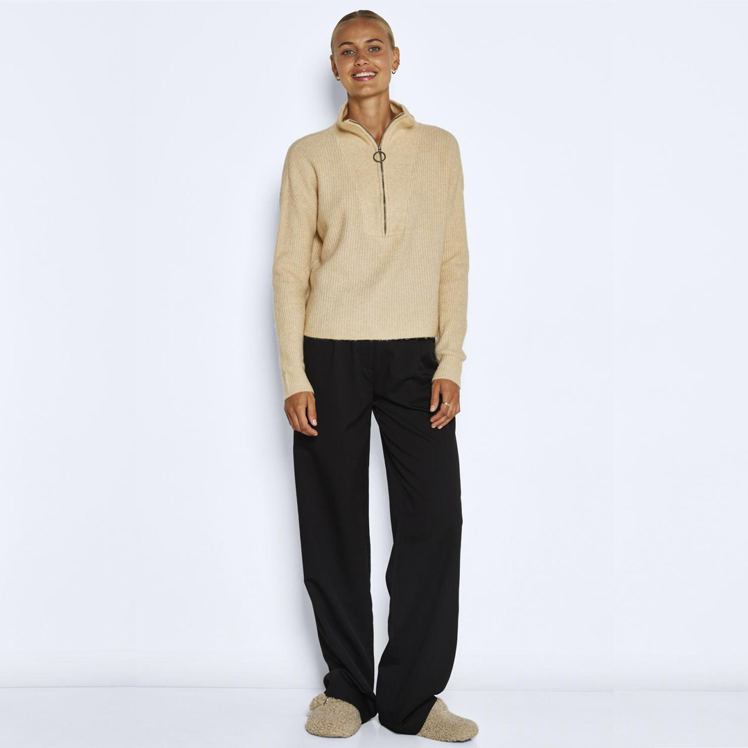 Mulher com camisola bege de malha com fecho na gola e calças pretas largas