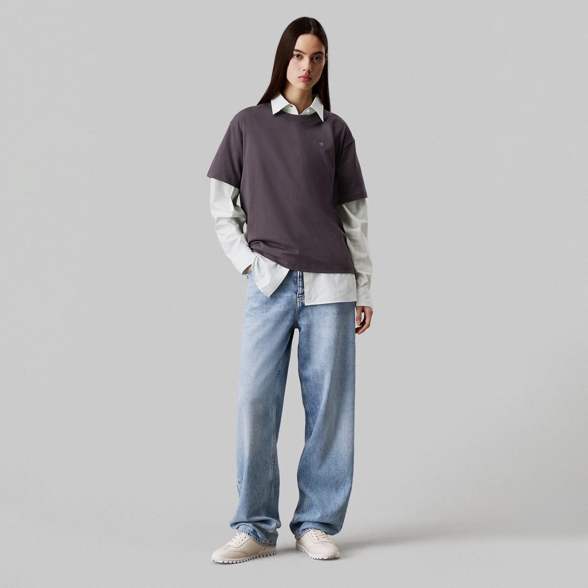 Modelo com t-shirt castanha escura, camisa branca, jeans azuis largos e ténis bege