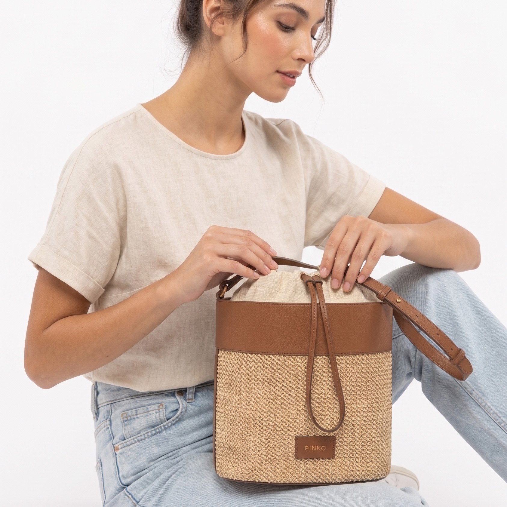 Mala tipo saco bege e castanha com detalhes em pele e etiqueta frontal PINKO segurada por mulher com t-shirt bege e jeans.