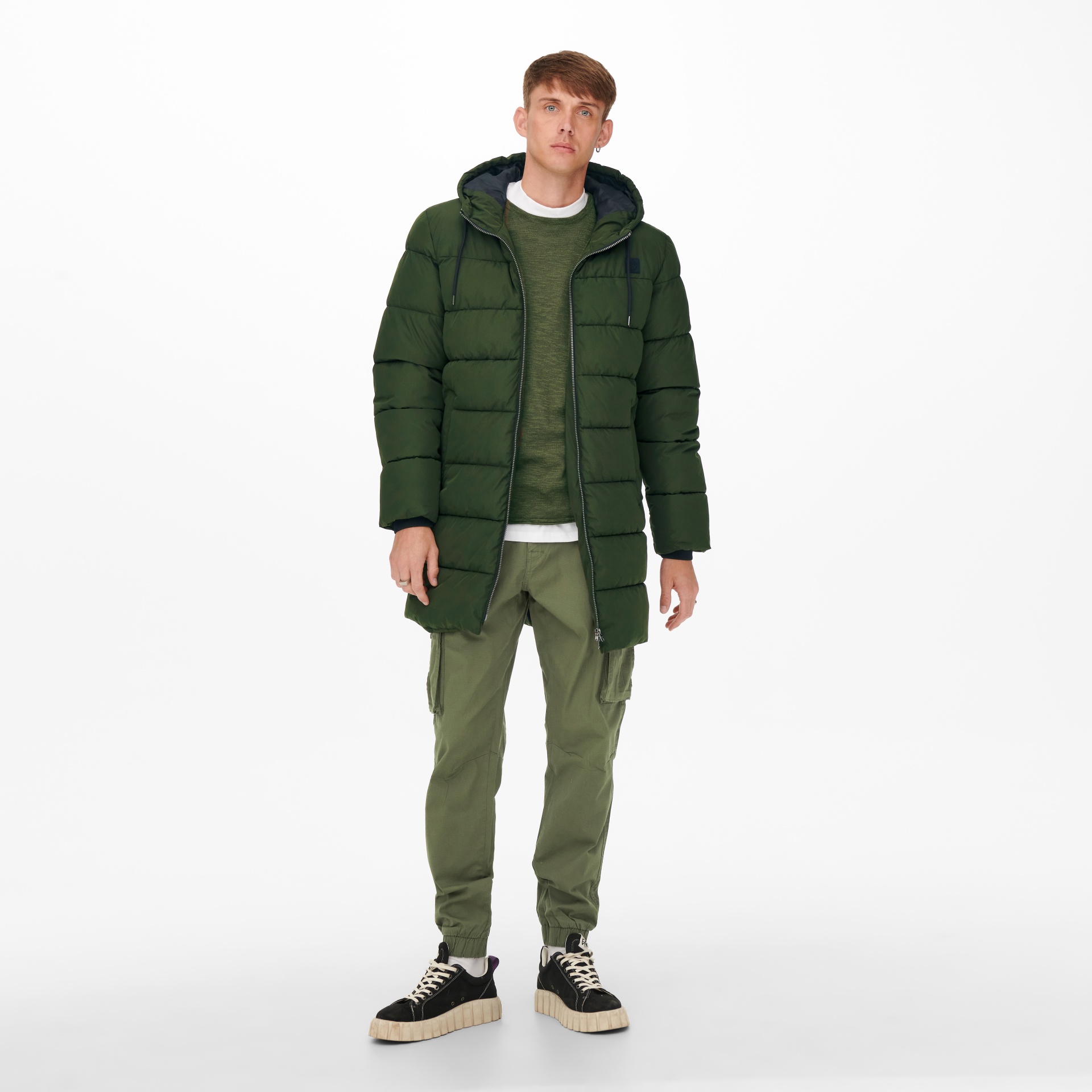 Homem com casaco acolchoado verde, camisola verde, calças cargo verdes e ténis pretos em fundo branco