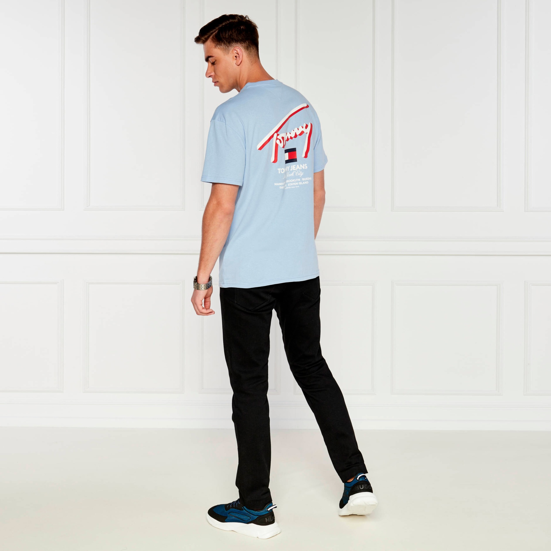 Homem com t-shirt azul claro com texto atrás, calças pretas e ténis azuis e pretos