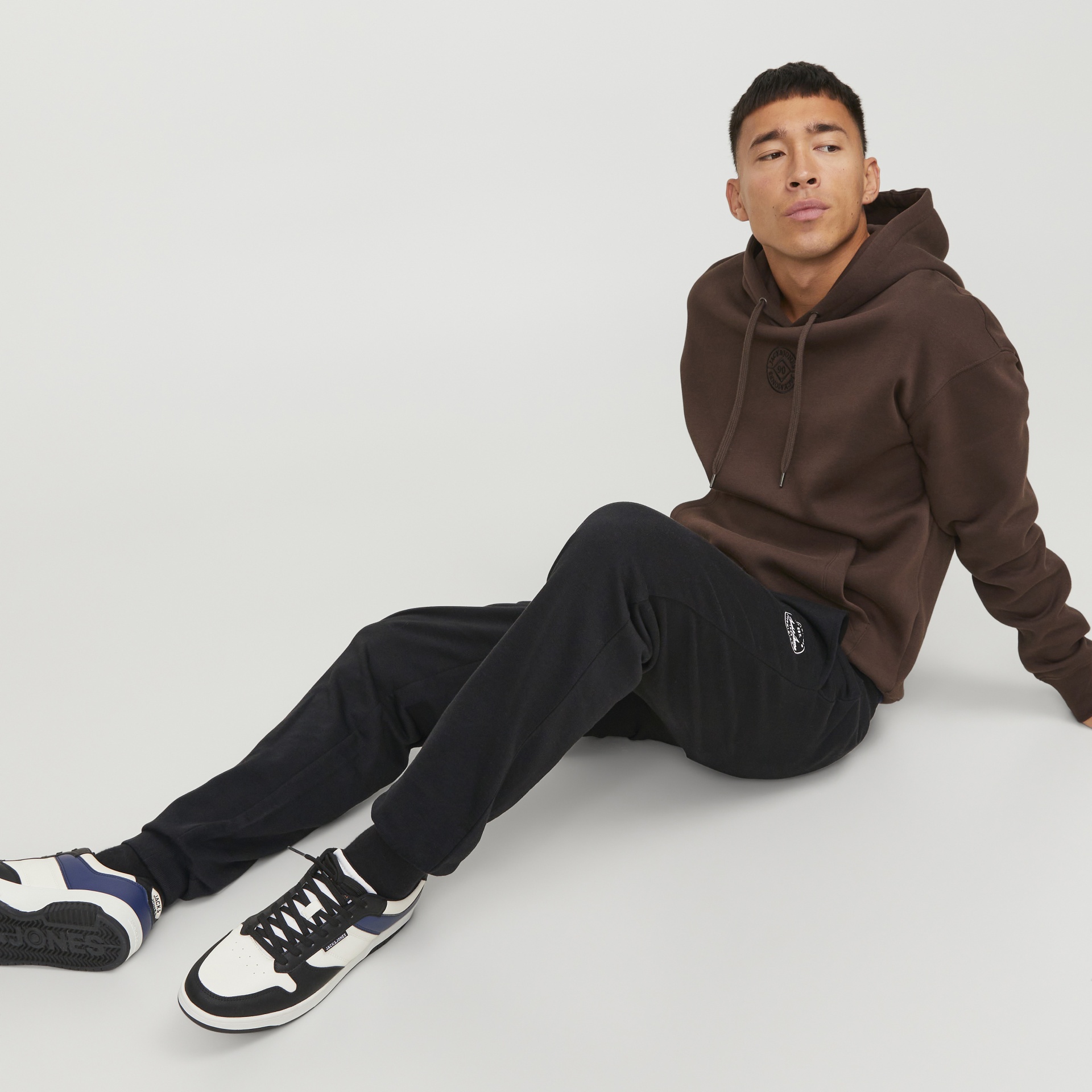Homem sentado com hoodie castanho escuro e calças pretas em fundo branco