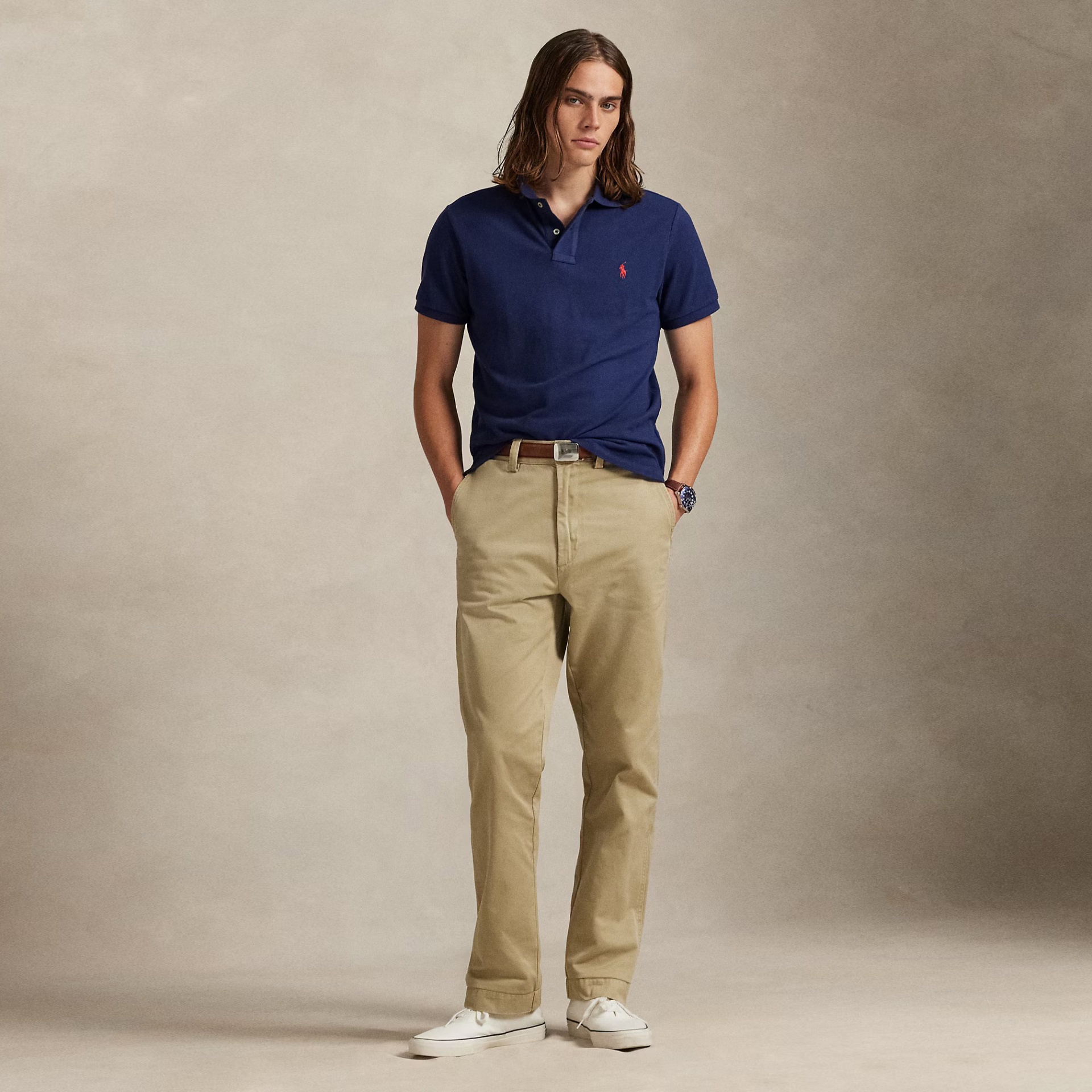Homem a usar polo azul escuro, calças bege e sapatilhas brancas, em fundo neutro.
