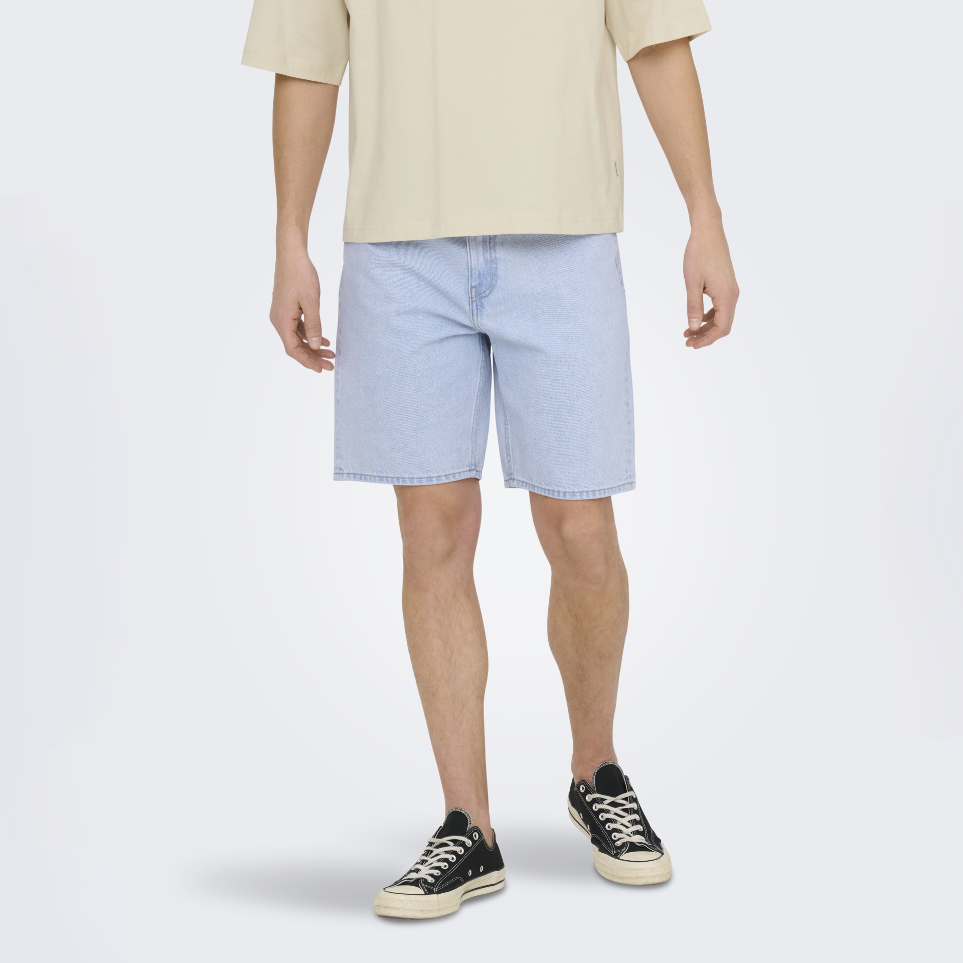 Homem de pé com t-shirt bege, calções de ganga azuis e ténis pretos.